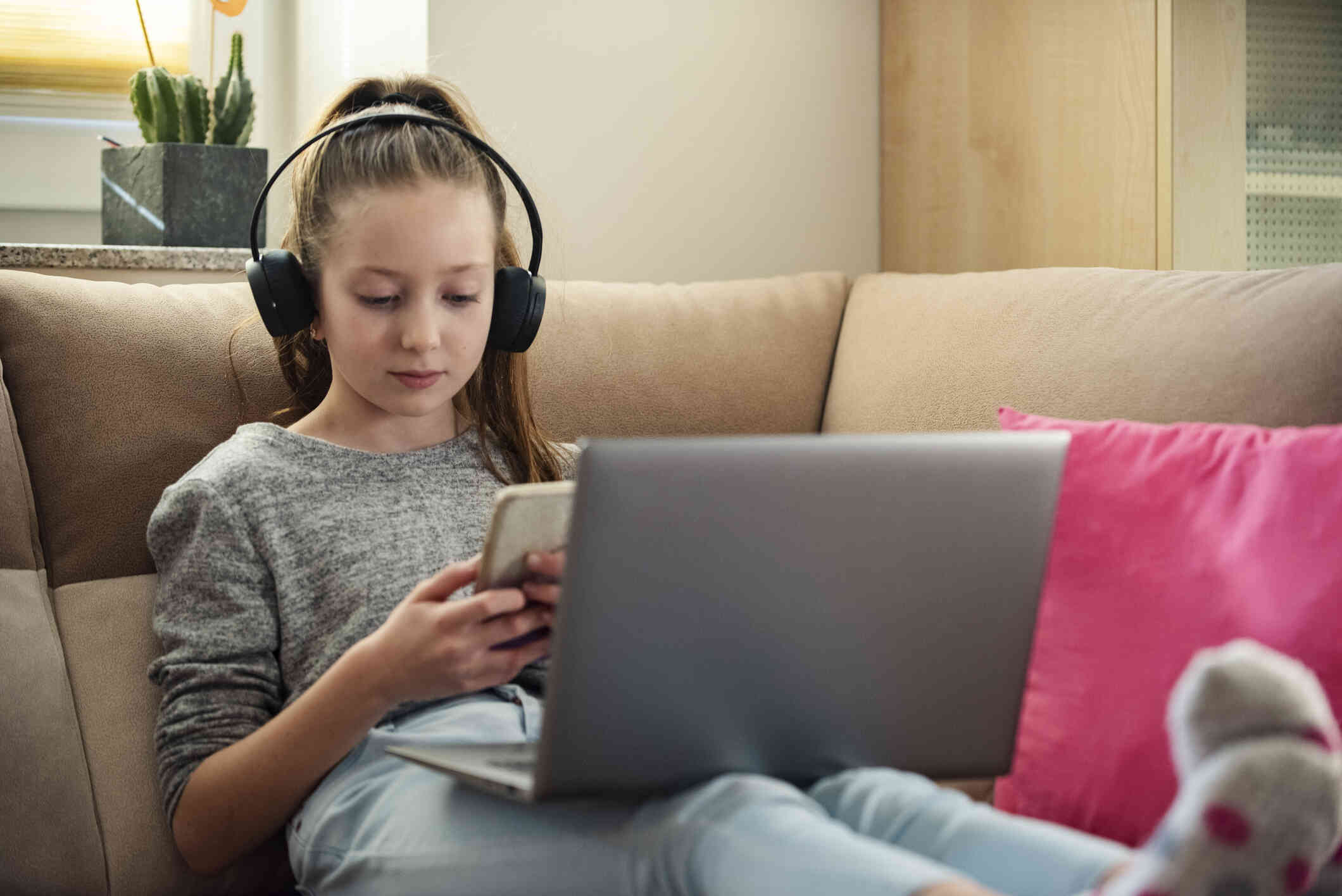 A teen girl reclines on the couch with with black headphones and her laptop open in her lap while looking down at the cellphone in her hand with a serious expression.